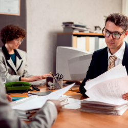 Mesa de madeira com uma caderneta com lápis de escrever e um borracha ao lado, e outro caderno maior com alguns textos na linguagem português e post-its colocado na borda